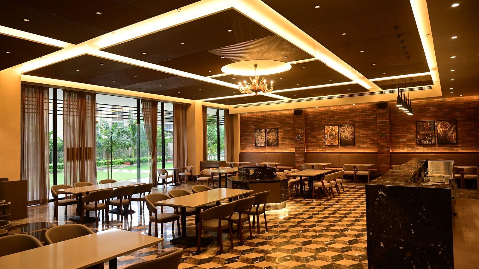 Dining area with tables, chairs, patterned floor, and textured walls, lit by ceiling lights and a chandelier, with a view to outside.