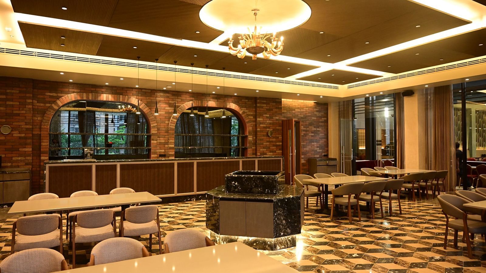 Dining room with tables, chairs, patterned floor, and textured walls, lit by ceiling lights and a chandelier, with a counter in the background.