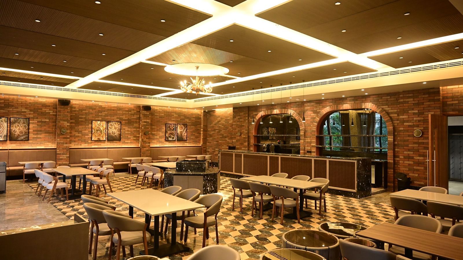 Dining area with multiple tables and chairs, patterned floor, and textured walls, lit by ceiling lights and a chandelier.