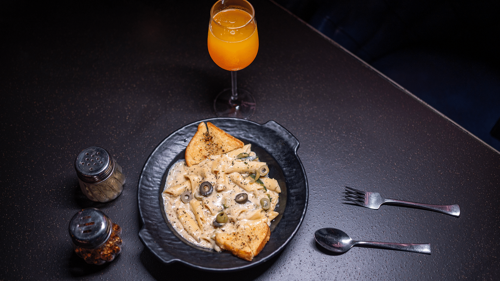 A top view of a plate of food garnished with cheese and accompanied by seasoning beside the plate, along with cutlery and a glass of drink, all set at Hotel Moti Palace