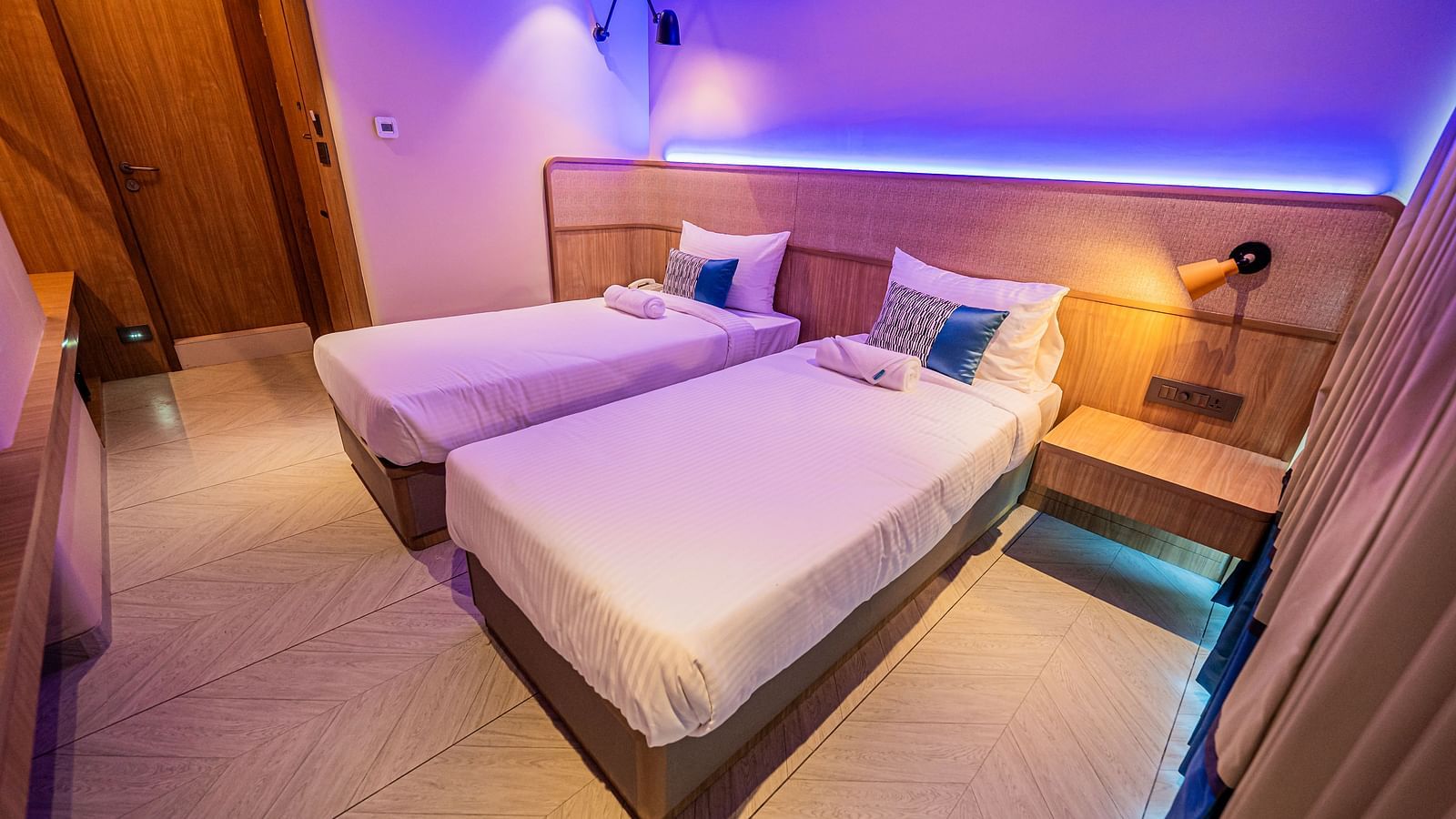 A spacious room at Indie Stays Mumbai International Airport, featuring two single beds with attached wooden headboards and cool lighting.