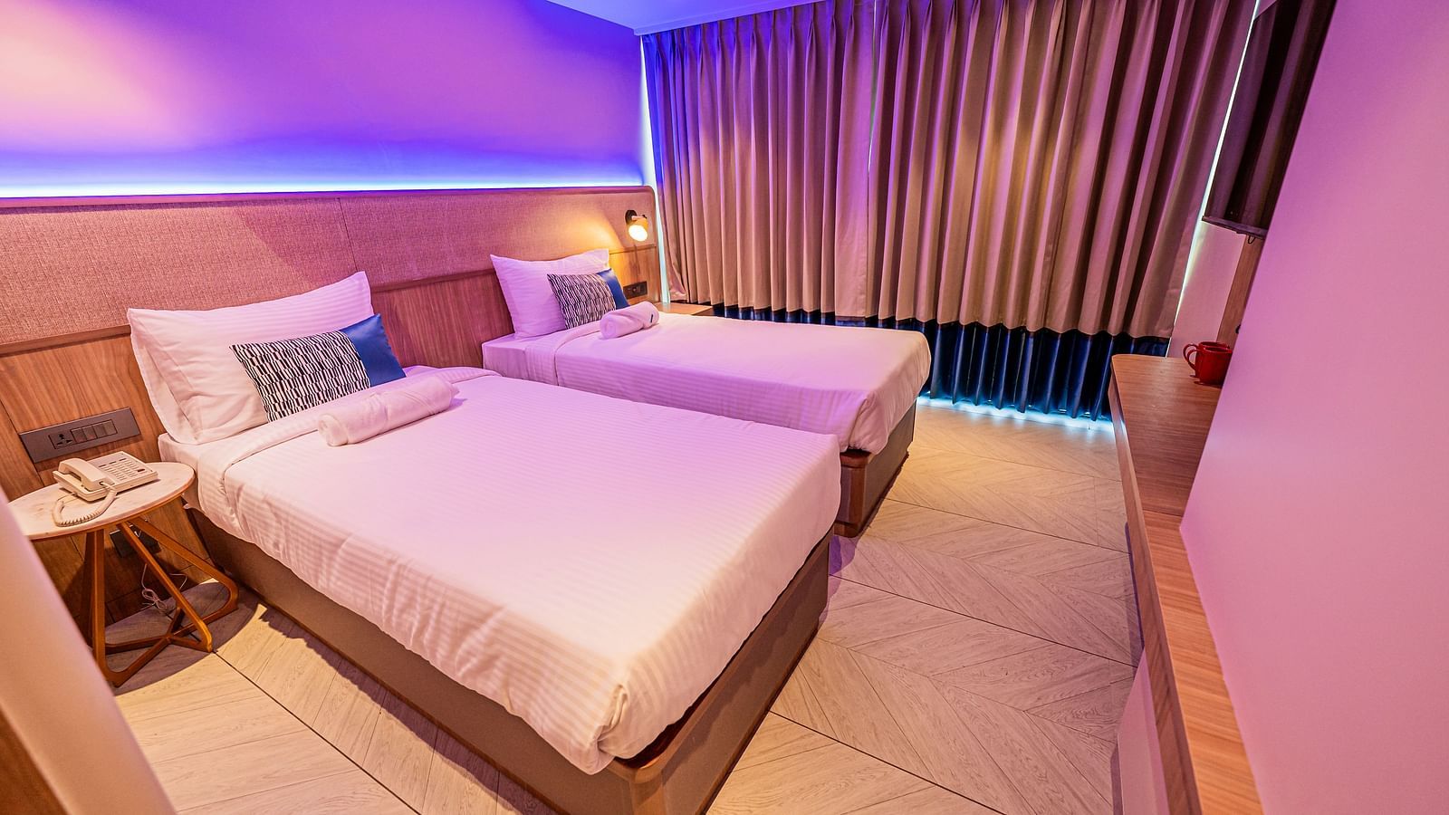 A side view of a room at Indie Stays Mumbai International Airport, featuring two single beds with attached headboards and windows fully covered with curtains.