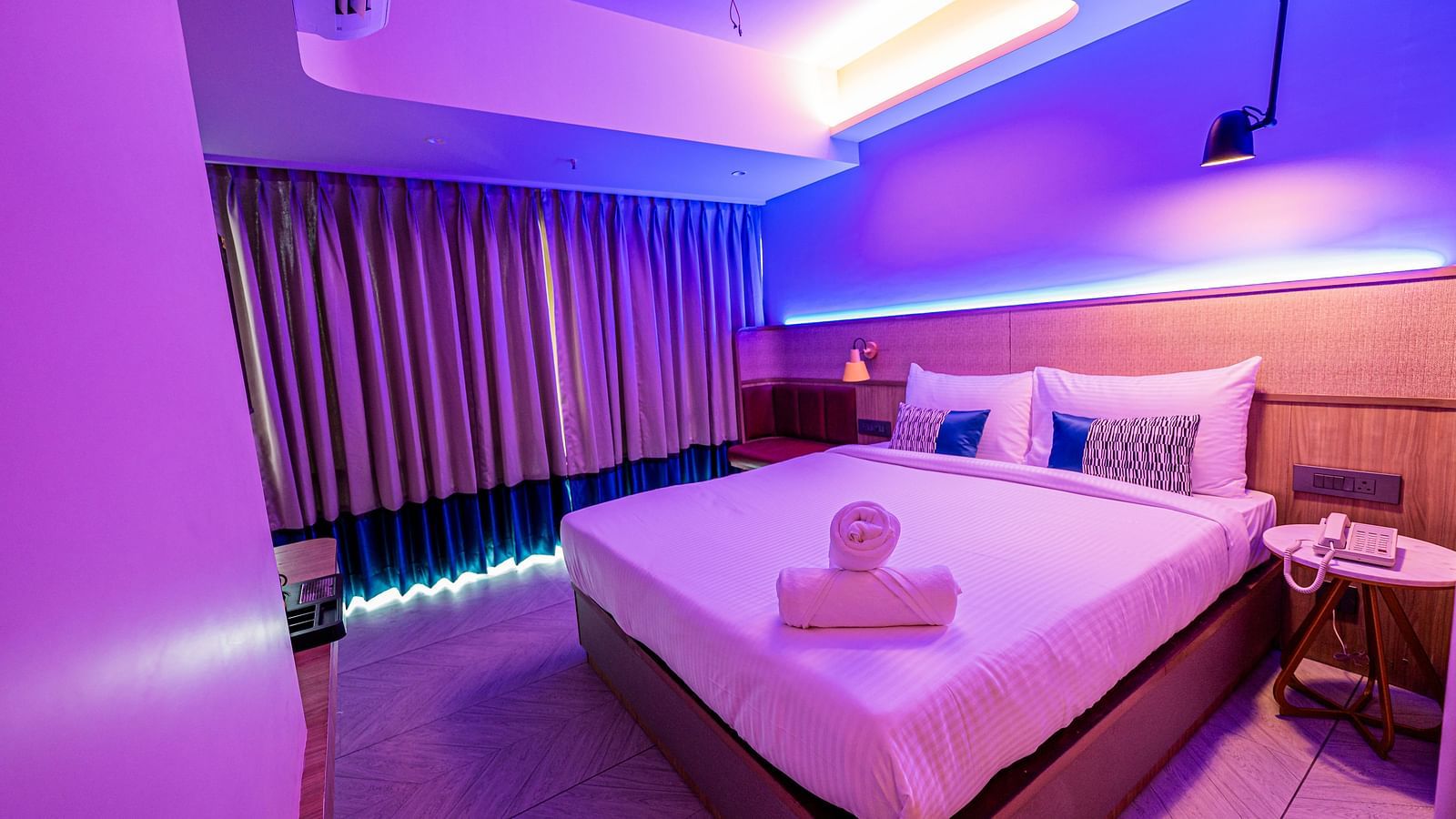 A spacious room at Indie Stays Mumbai International Airport features a large bed with neatly rolled towels, a window with closed curtains, and the room lit with cool lighting.