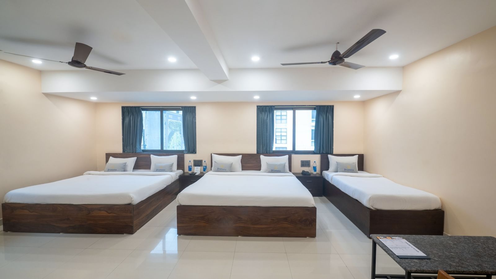 A large room at Pravasa Stay, Shirdi, contains three beds, two ceiling fans, and multiple windows.