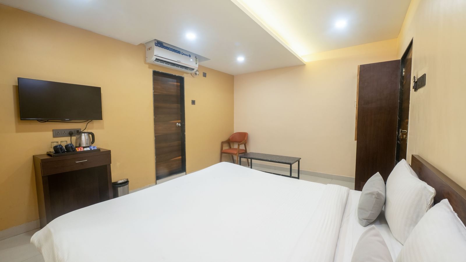 A room at Pravasa Stay, Shirdi, features a large bed with white linen, a desk, and a wall-mounted television.
