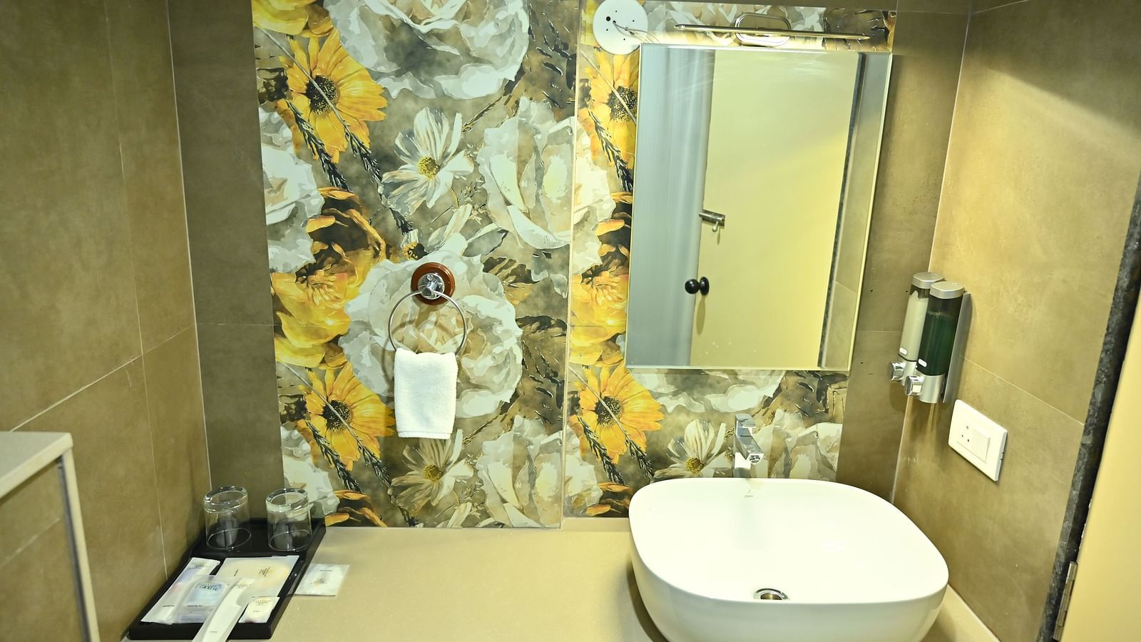 A well-lit bathroom at TGI Livancia Hillside, JC Nagar, Mysore, featuring a modern sink, a mirror with decorative floral tiling, and provided toiletries.