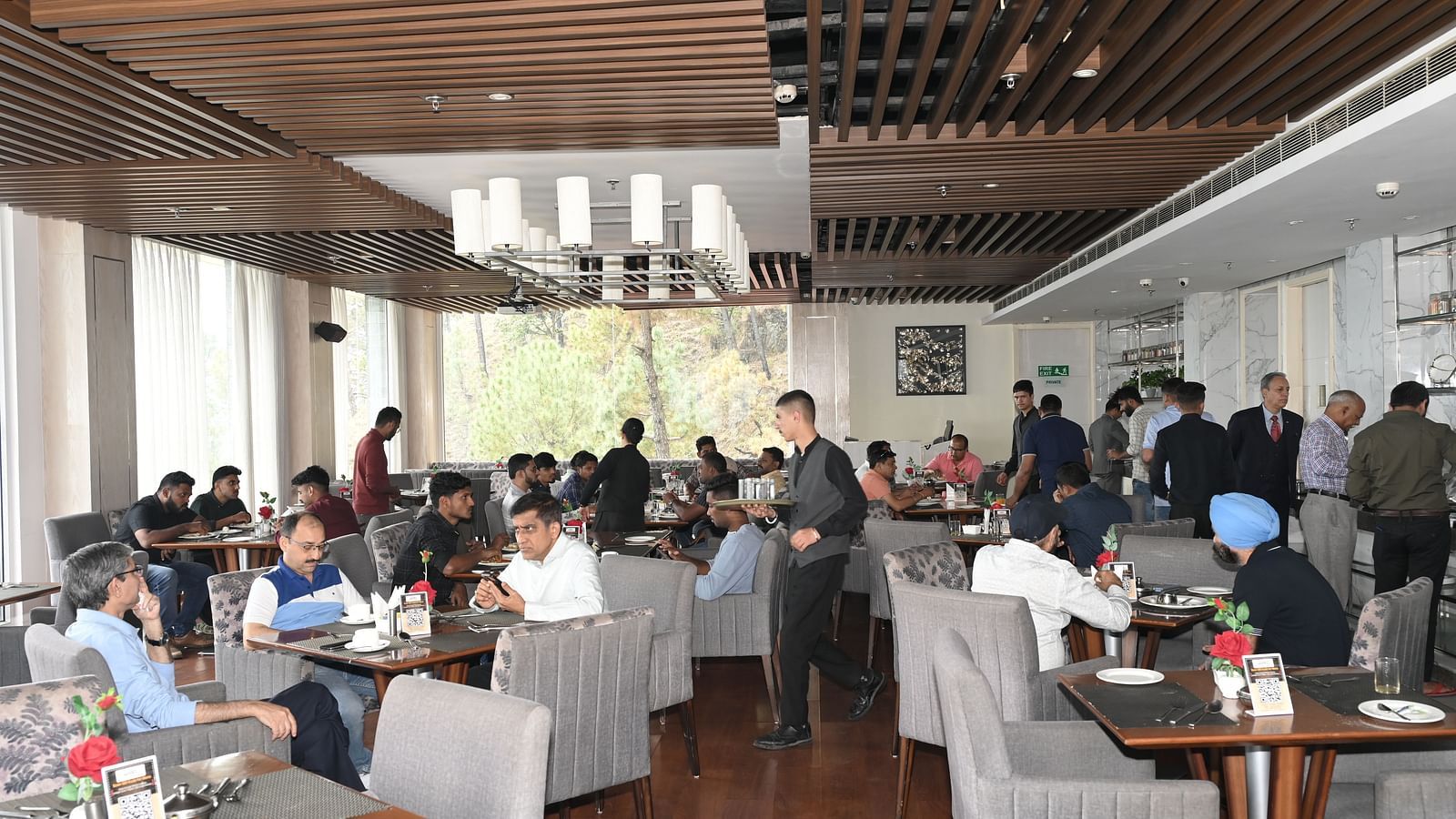 A lively and elegant restaurant setting with Refex team members and guests engaged in conversation and dining, as servers move around efficiently in a warm, well-lit atmosphere with modern wooden interiors - The Zion, Shimla.