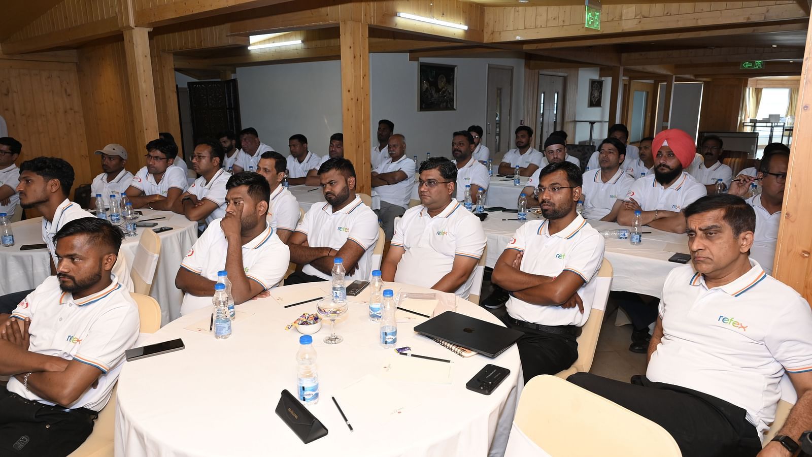 Group of professionals in white 'Refex' shirts attentively participating in a conference or training session in a wooden-panelled meeting room - The Zion, Shimla.