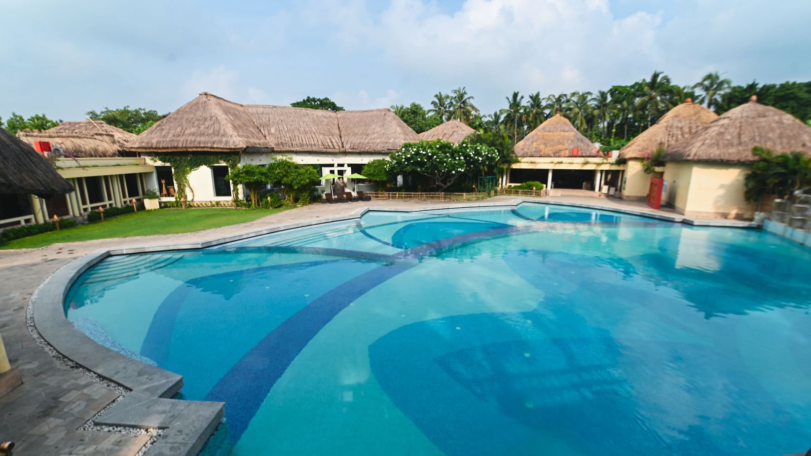 A daylight view of the common swimming pool at Vedic Village Spa Resort, featuring buildings with tatched roofs in the background.