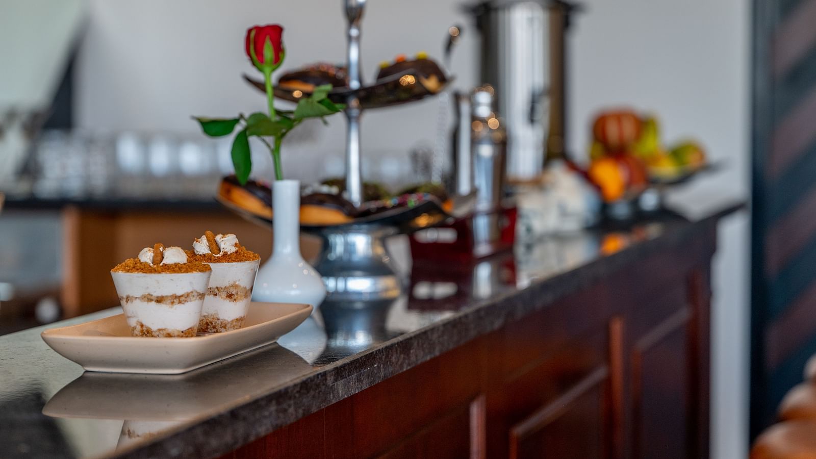 A dark wooden counter with a marble top holding various items, including a tray with two small desserts, a vase with a red rose and other bottles. - Vishranti Hotel, Dehradun.