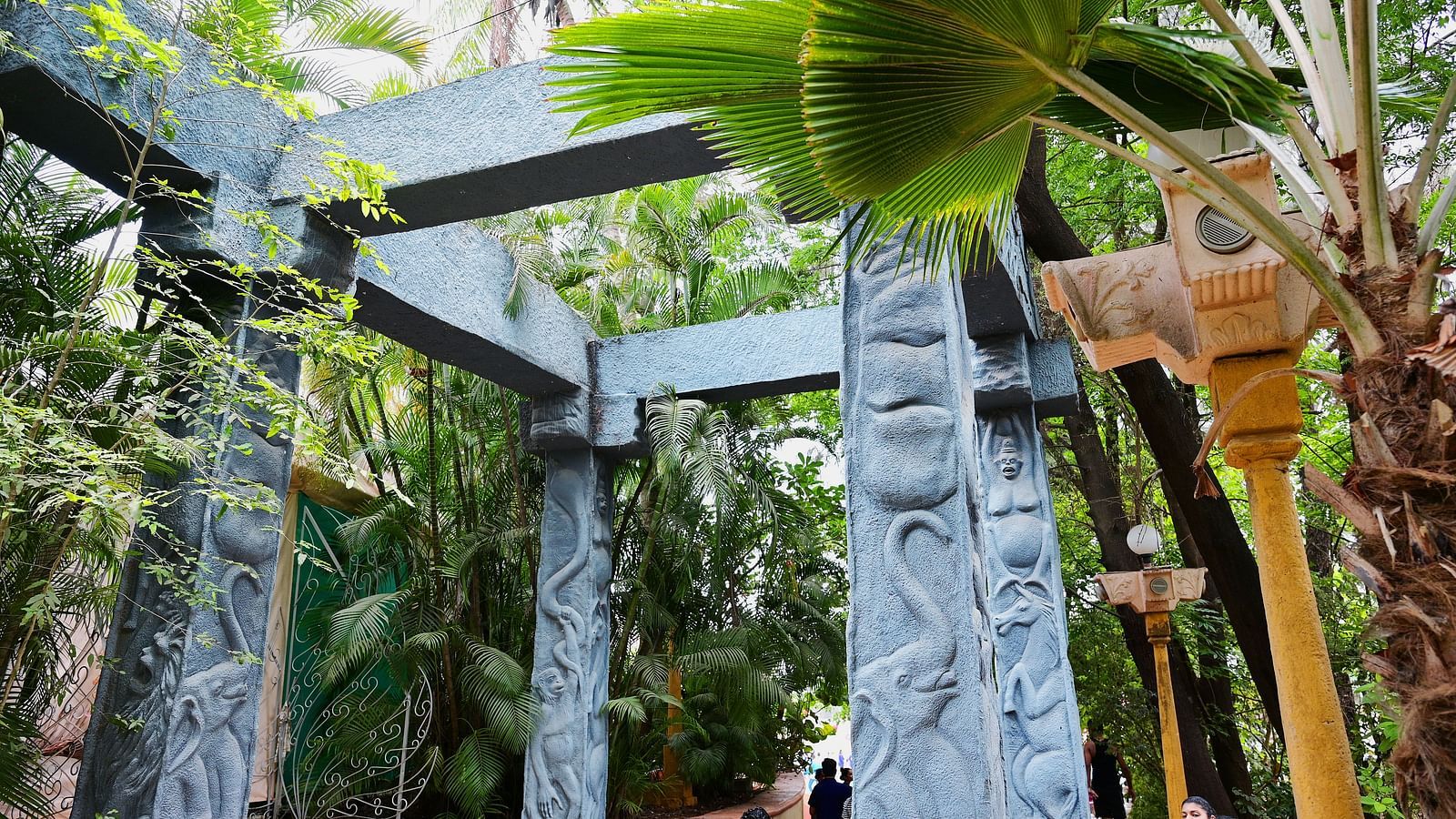 Wooden pergola structure with hanging plants and a stone pathway in a tropical garden.