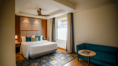 A brightly lit hotel room features a neatly made bed with white linens, a teal armchair, and a small round side table with a lamp. A window with sheer curtains allows natural light to filter in - Ananta Spa and Resort, Pushkar