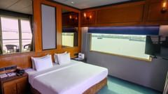 A hotel bedroom with a large bed and a wide window offering a view of the river. Wooden paneling and a TV are visible.