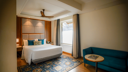 A brightly lit hotel room features a neatly made bed with white linens, a teal armchair, and a small round side table with a lamp. A window with sheer curtains allows natural light to filter in - Ananta Spa and Resort, Pushkar
