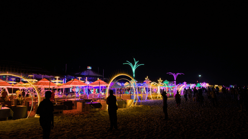 A dark beach at night is illuminated by colourful string lights and unique glowing structures, with several people visible in the distance | Christmas Carnival Goa