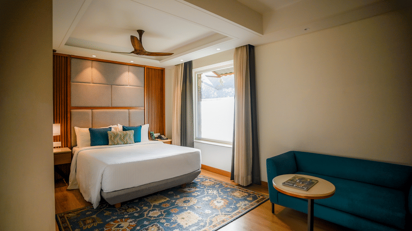 A brightly lit hotel room features a neatly made bed with white linens, a teal armchair, and a small round side table with a lamp. A window with sheer curtains allows natural light to filter in - Ananta Spa and Resort, Pushkar
