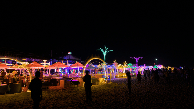 A dark beach at night is illuminated by colourful string lights and unique glowing structures, with several people visible in the distance | Christmas Carnival Goa