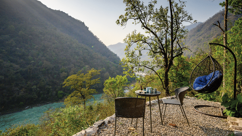A scenic outdoor seating area featuring a small table with chairs and a purple hanging nest chair overlooking the turquoise waters of the river at Neemrana’s Glasshouse on The Ganges.