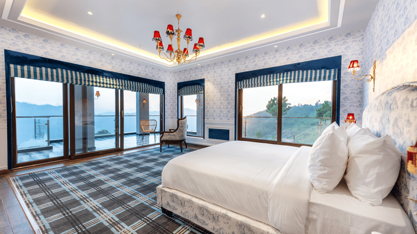 A bright, elegant interior of the Presidential Suite at The Manor Sports & Wellness Hotel featuring a large white bed, plaid carpet, statement chandelier, and floor-to-ceiling windows with mountain and lake views.