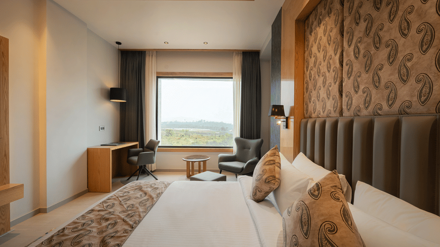A bright guest room with a king-sized bed, a patterned headboard, and a large window overlooking the hills at Ananta Express, Katra.