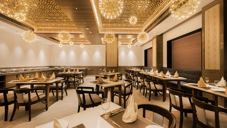 A spacious restaurant dining hall featuring wooden tables, black chairs, and modern gold-tone chandeliers at Ananta Express, Katra.