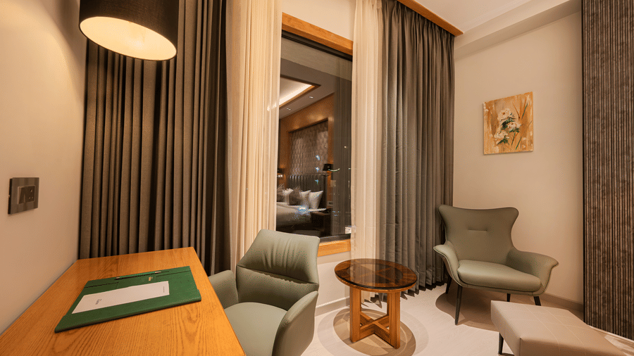 A bedroom corner featuring a wooden desk, a green armchair, and floor-to-ceiling grey curtains at Ananta Express, Katra.