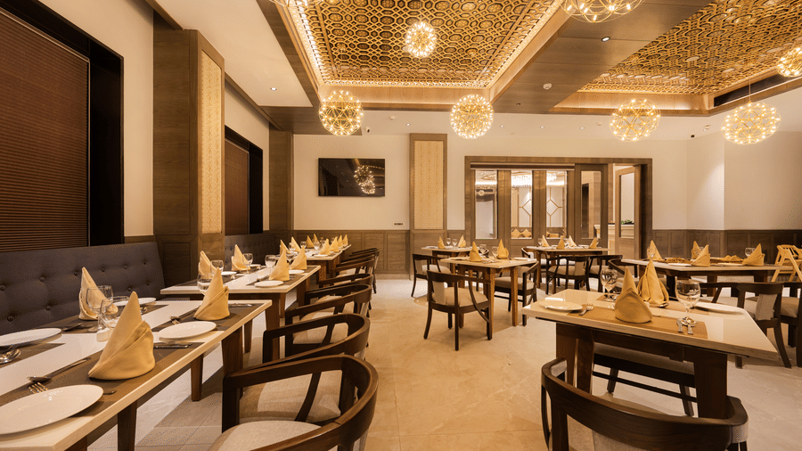A well-lit dining room with rows of wooden tables and chairs under an elegant decorative ceiling at Ananta Express, Katra.
