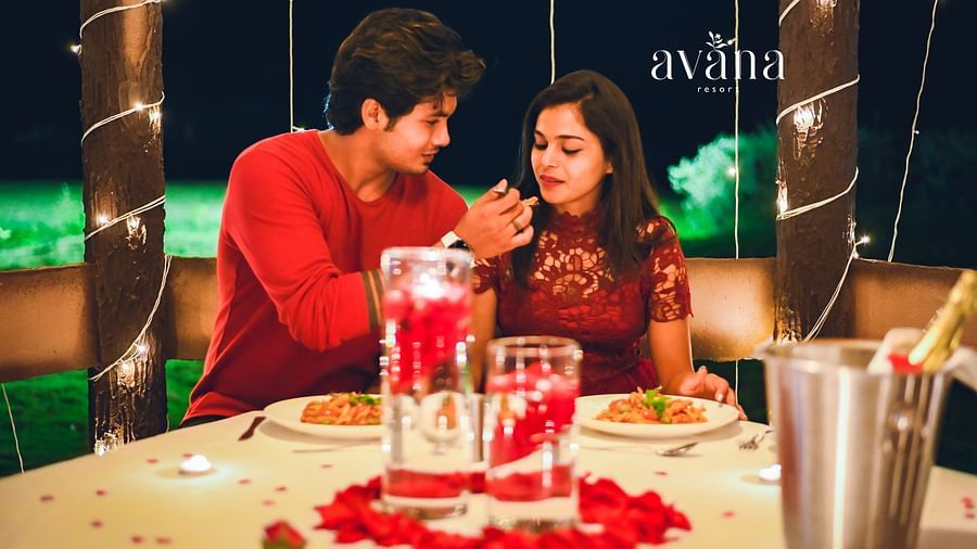 A couple enjoying a romantic dinner with a heart-shaped rose petal arrangement on the table, champagne, and candles.