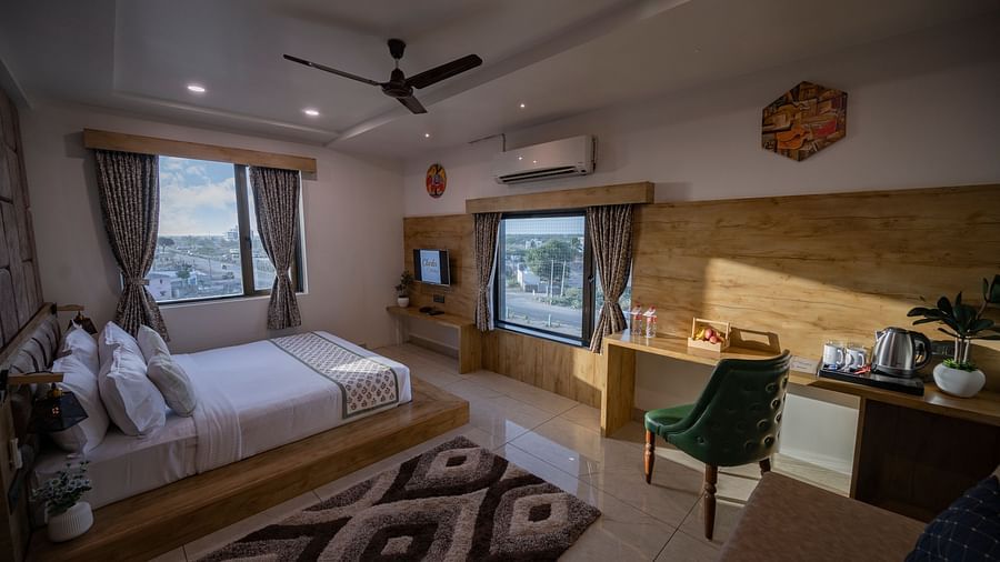 A room with double bed with white linens, chair, large window, desk, and wall-mounted TV at Clarks Collection Somnath.