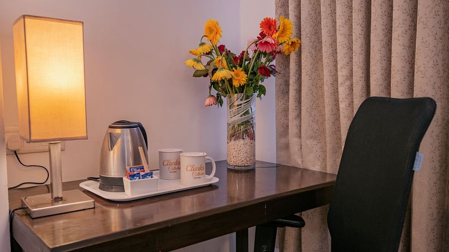 Close-up of a desk with a lamp, kettle, two cups, tea sachets, and a vase of flowers near curtains at Clarks Collection, Vadodara.