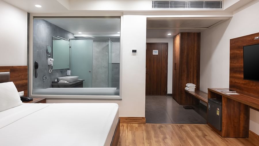 A modern hotel room with a large bed, wooden flooring, a TV, and a glass-walled bathroom in view.- Clarks Inn Express, Near Jammu Bari Brahmana Railway Station