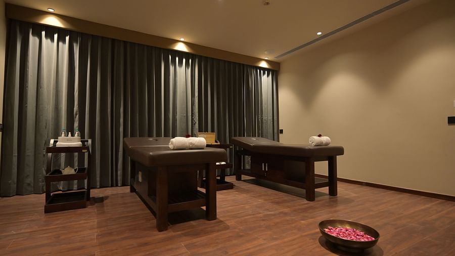 A softly lit spa treatment room features two massage tables with rolled towels, dark curtains, and a bowl of rose petals on the floor. - Clarks Safari Haridwar