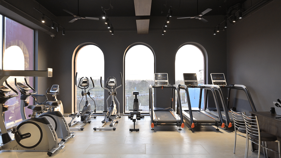 A well-equipped gym featuring cardio machines like treadmills and elliptical, along with stationary bikes and weight training equipment, set against a backdrop of arched windows and dark walls. - Clarks Safari Haridwar