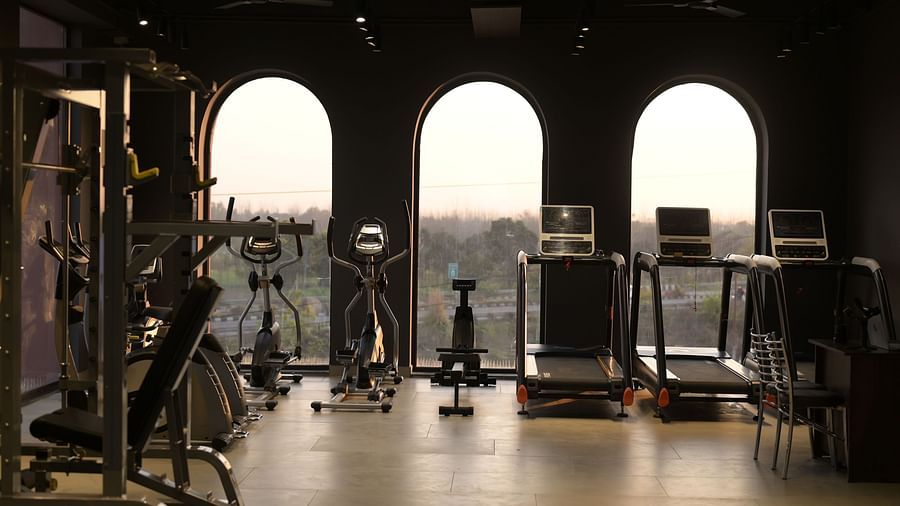 A well-equipped gym with various exercise machines, including treadmills and weightlifting equipment, set against a backdrop of arched windows overlooking a cityscape. - Clarks Safari Haridwar