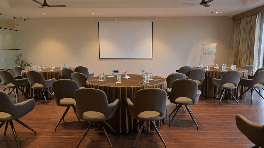 A meeting room set up with round tables and chairs, a projector screen, and ceiling fans. - Clarks Safari Haridwar