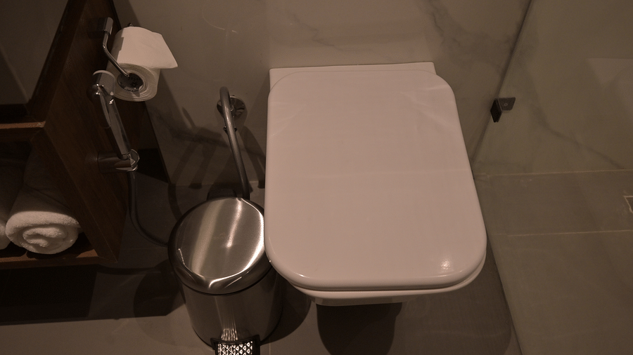 A top-down view of a closed, white toilet with a silver trash can next to it and a toilet paper holder on the wall in Deluxe Room. - Clarks Safari Haridwar