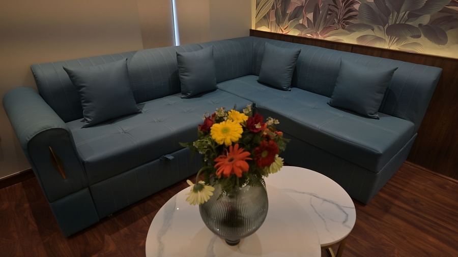 A sectional sofa with blue cushions is positioned in Premium Room, with a white kidney-shaped table and a vase of flowers in front of it. -  Clarks Safari Haridwar