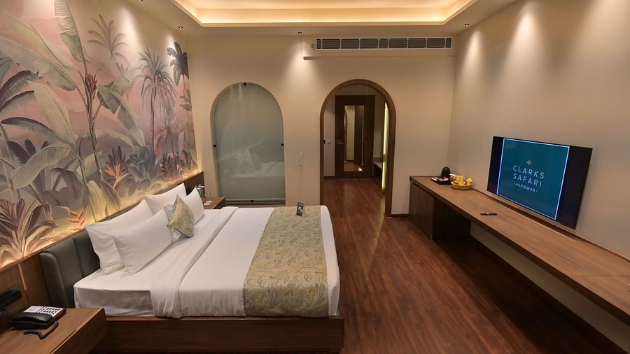 Premium Room featuring a king size bed with white linens and decorative pillows, a mural of a tropical landscape, an arched doorway, a long wooden desk with a TV, and hardwood floors. - Clarks Safari Haridwar