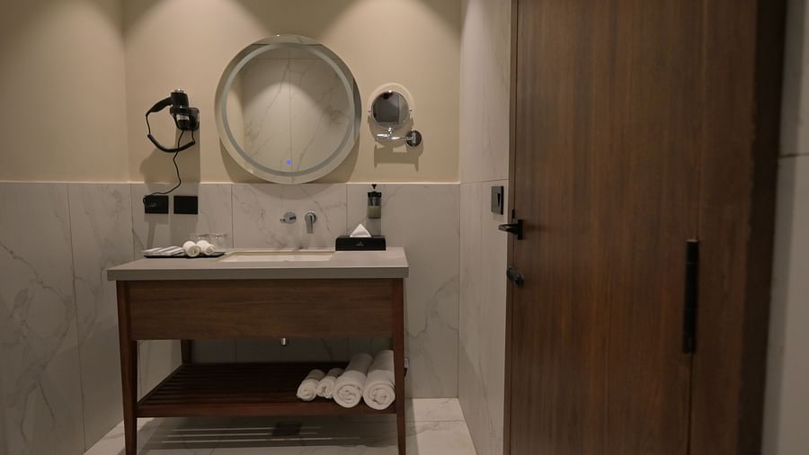 A bathroom vanity area with a wooden cabinet, a round mirror, and decorative items, along with a closed wooden door on the right of Premium Room. - Clarks Safari Haridwar