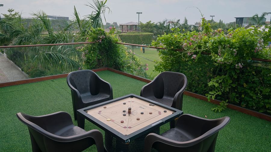 An outdoor seating area at Clarks Safari Kheda with a table, four chairs, and a board game set up, on a low,  surrounded by green surface and trees.