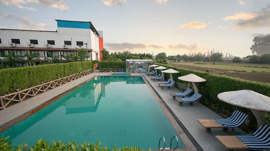 At Clarks Safari Kheda, a long, rectangular swimming pool bordered by lounge chairs under umbrellas, with the hotel building and fields nearby.