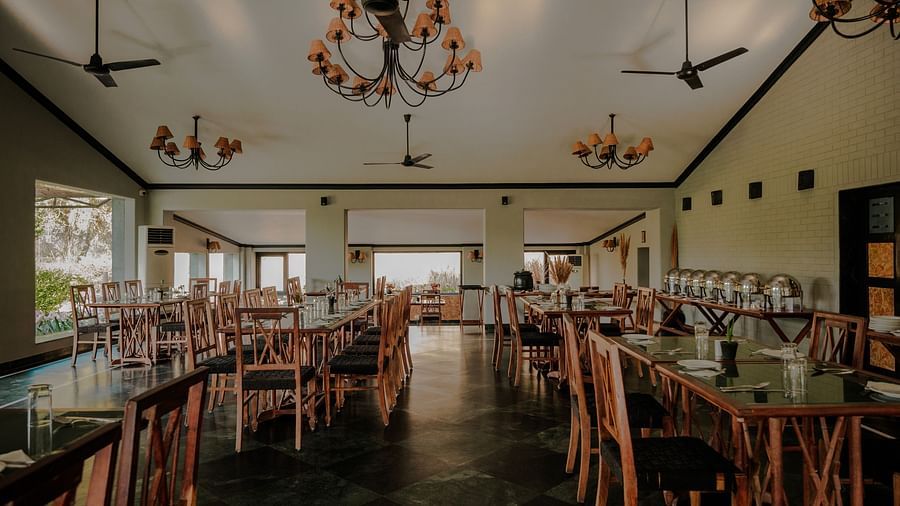 Interior of a vacant dining room with multiple wooden tables and chairs arranged in rows. Floral wreath chandeliers illuminate the space. Windows are on the left - Clarks Safari, Puskhar