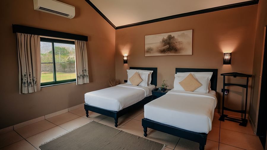 Hotel room with two single beds, brown walls, a window with curtains, and an air conditioning unit. A rug sits on the tiled floor - Clarks Safari, Pushkar