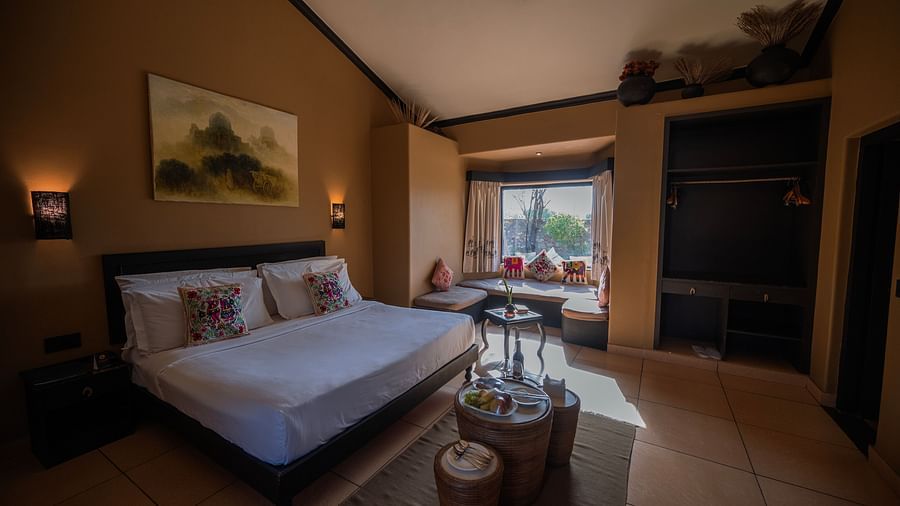 Interior of a hotel room showcasing a double bed, brown walls, a painting, and a window seat area with pillows and a table - Clarks Safari, Pushkar