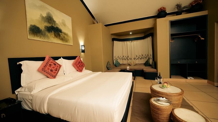 A plush bed with fresh linen with a wall hanging over its headboard and fruits and a wine bottle with a couple glasses in front of the bed in Superior Cottage at Clarks Safari Pushkar