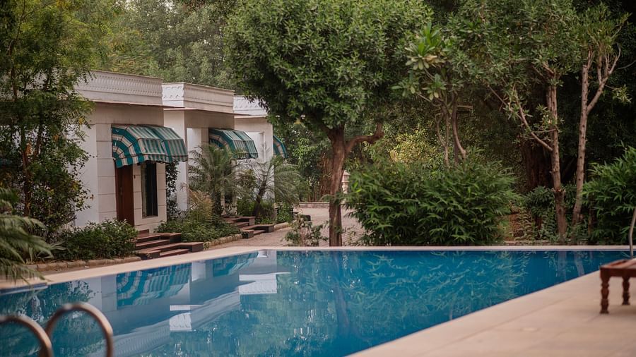 A rectangular swimming pool with clear blue water stretches towards a white building with green awnings, surrounded by trees - Clarks Safari, Ranthambore