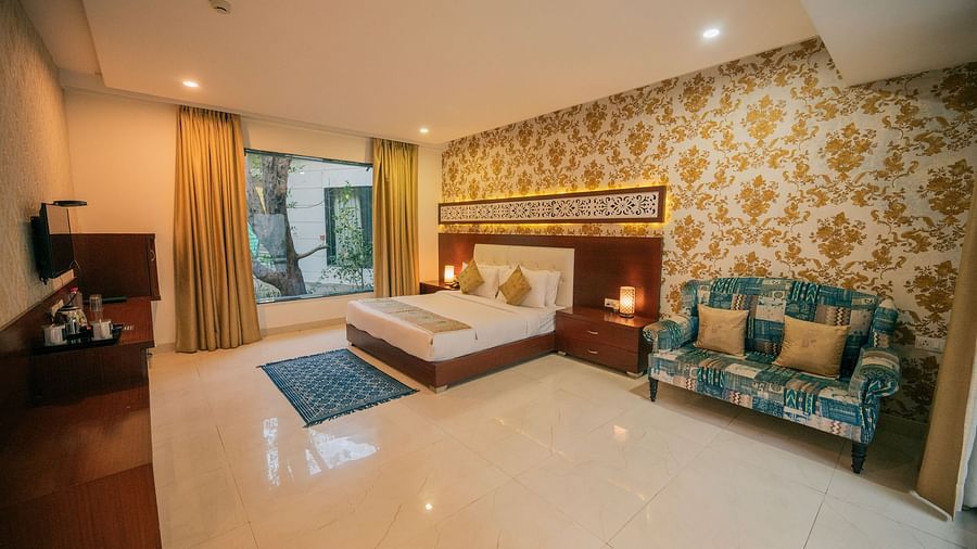 Wide view of a luxurious hotel room featuring a king-size bed, a stylish blue sofa, a dark wooden dresser, and large windows with sheer curtains - Clarks Safari, Ranthambore