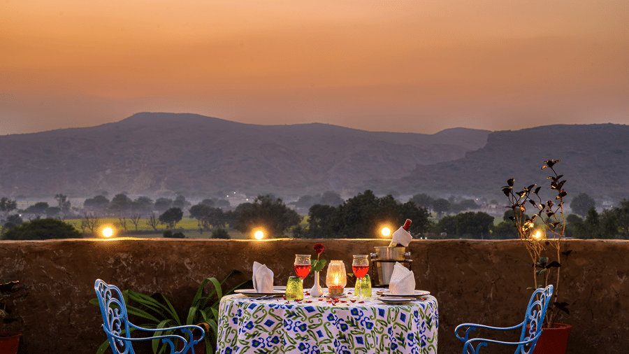 A blue patterned outdoor dining table set with drinks and napkins on an open-air terrace overlooking hills at sunset -  Hill Fort-Kesroli