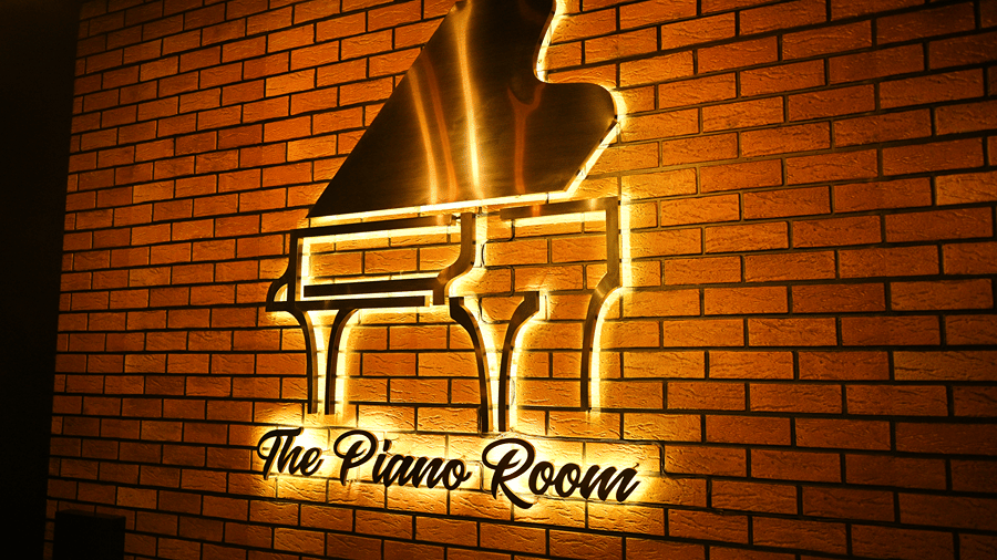 Backlit metal grand piano sign with the words The Piano Room mounted on an exposed red brick wall in the restaurant at HM Hotel & Resort, Dibrugarh.