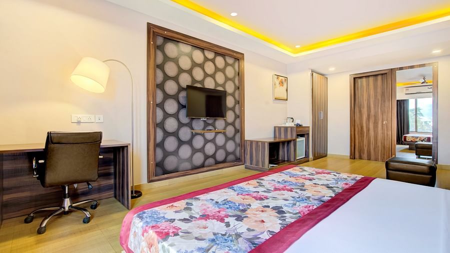 Bedroom featuring a double bed with floral bedding, study desk, TV unit, and soft ceiling lighting at Voyage Milestone Hotel and Banquet Siliguri